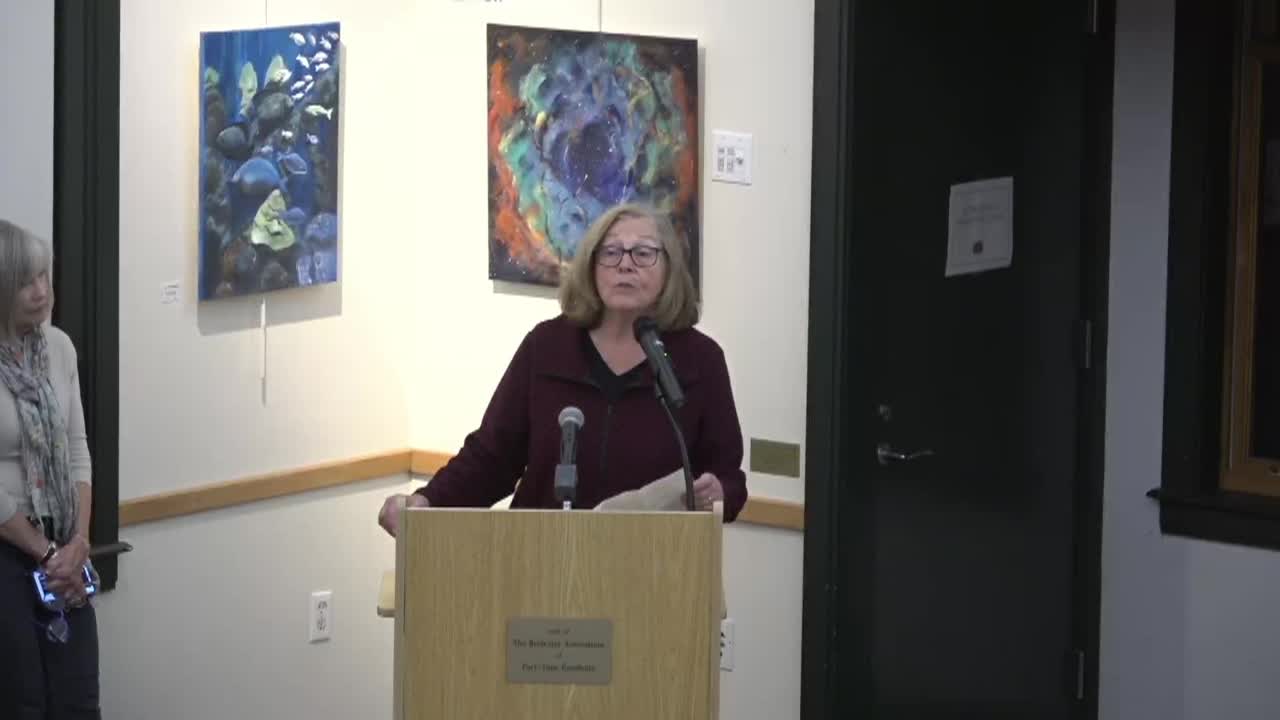 A person stands at a podium in a corner that has two abstract pieces of art hanging on the wall